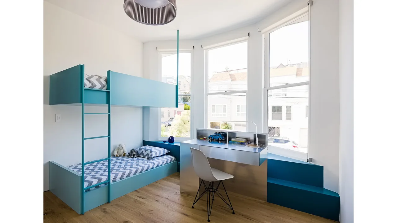 This bedroom includes blue bunk beds and a built-in blue desk facing three large windows that provide natural light.