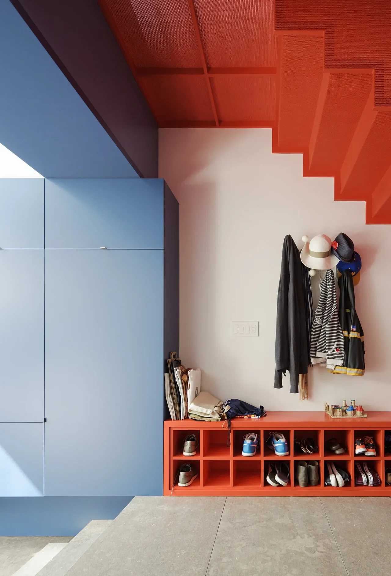 Entryway features bold blue cabinetry and a bright orange shoe cubby bench beneath a perforated orange staircase.