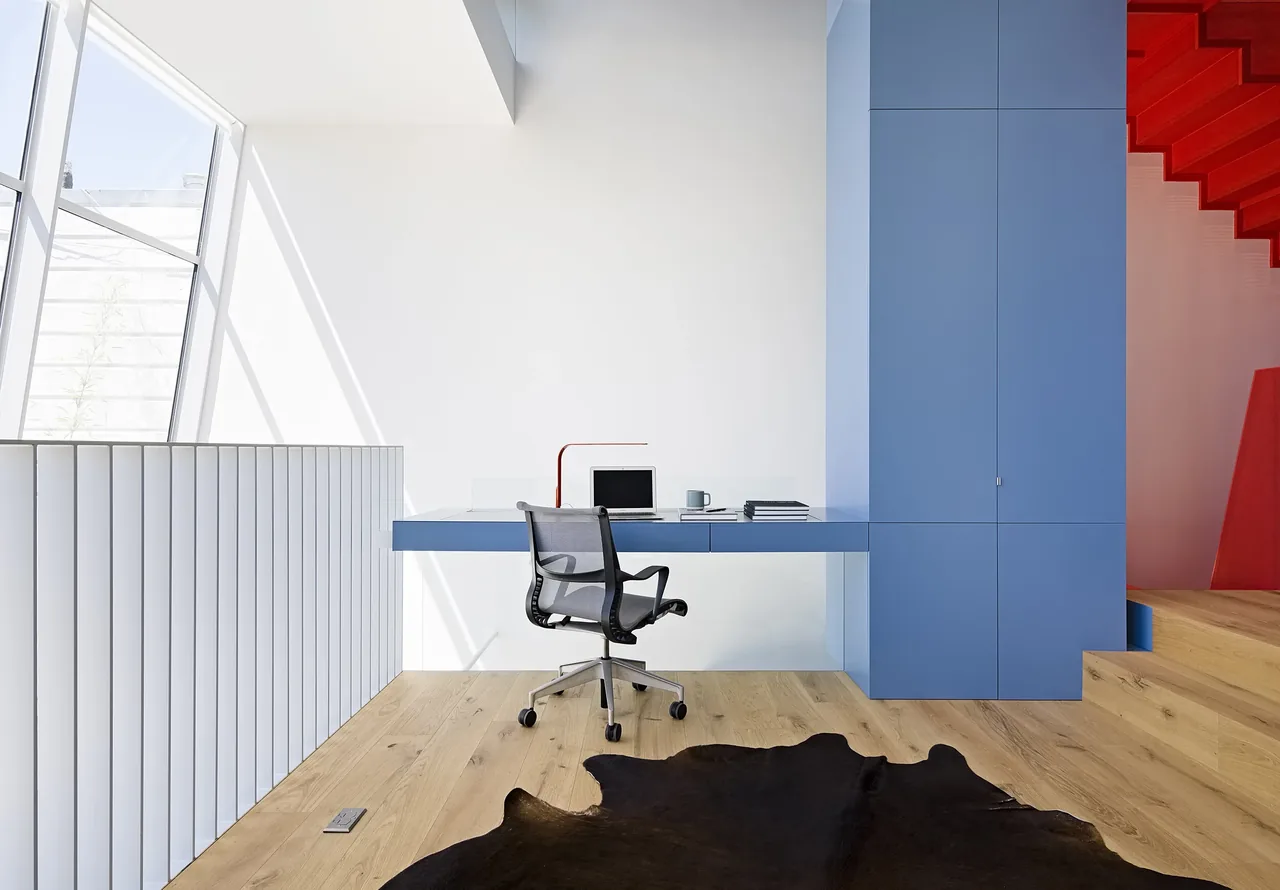 Home office space featuring a floating blue desk and black mesh chair with natural wood flooring and a red staircase.