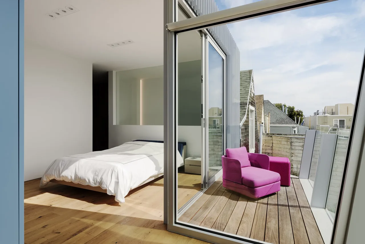 Modern bedroom features floor-to-ceiling sliding glass doors that lead to a balcony with pink seating and wood decking.