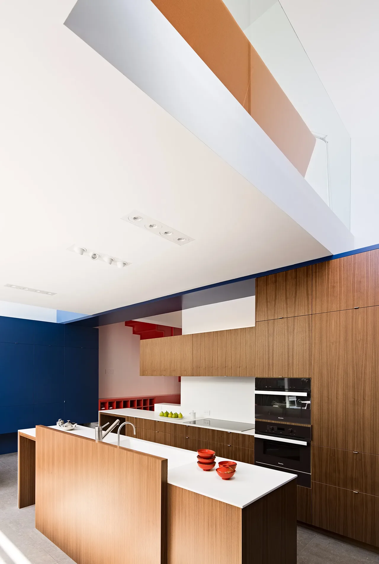 The CUT OUT HOUSE kitchen features built-in wood cabinetry, a white island countertop, and built-in ovens with colorful accents.