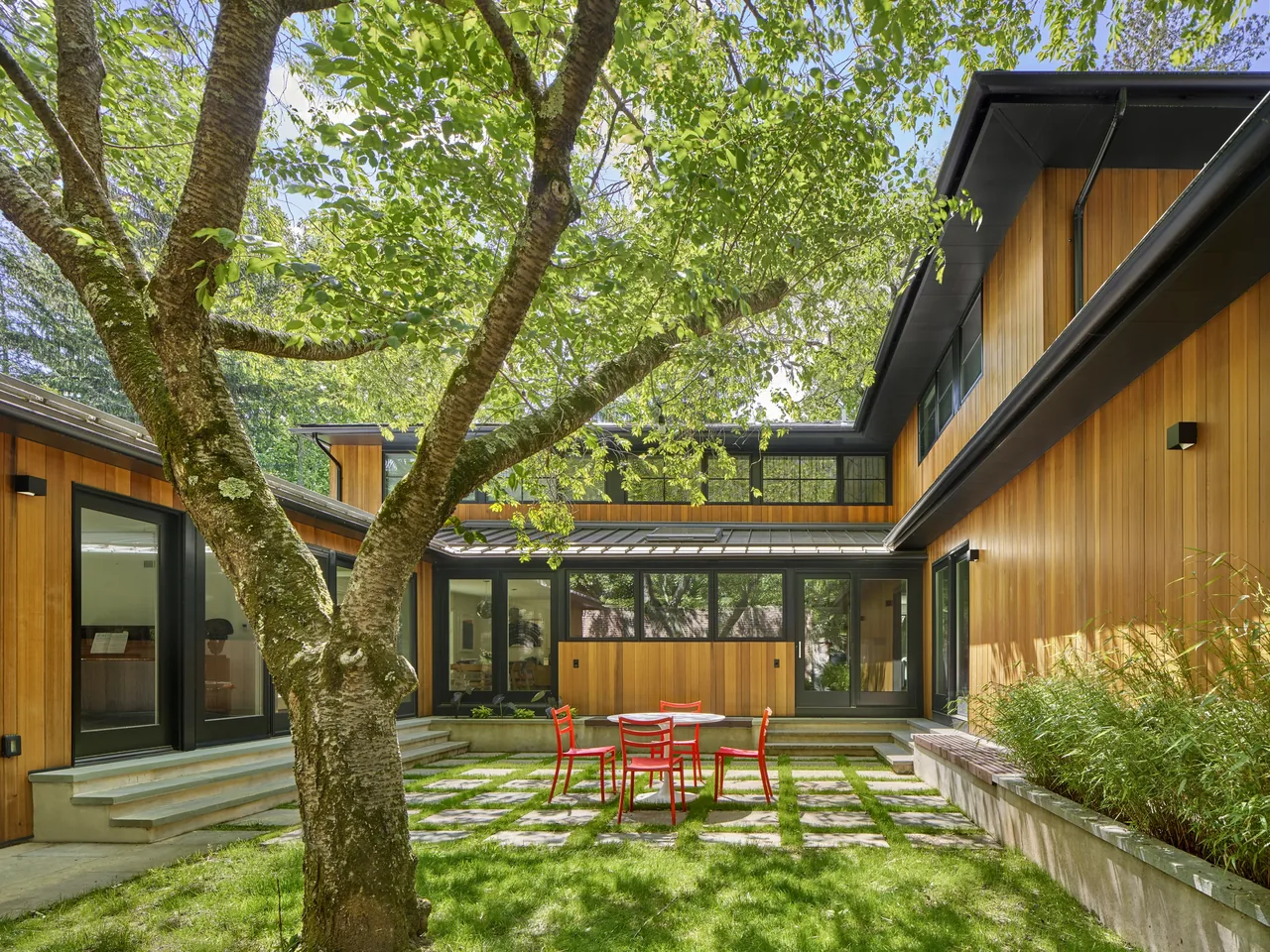 Rear view of a two-story house with wood siding and black window frames surrounding a patio with a tree, red chairs, and a metal table.