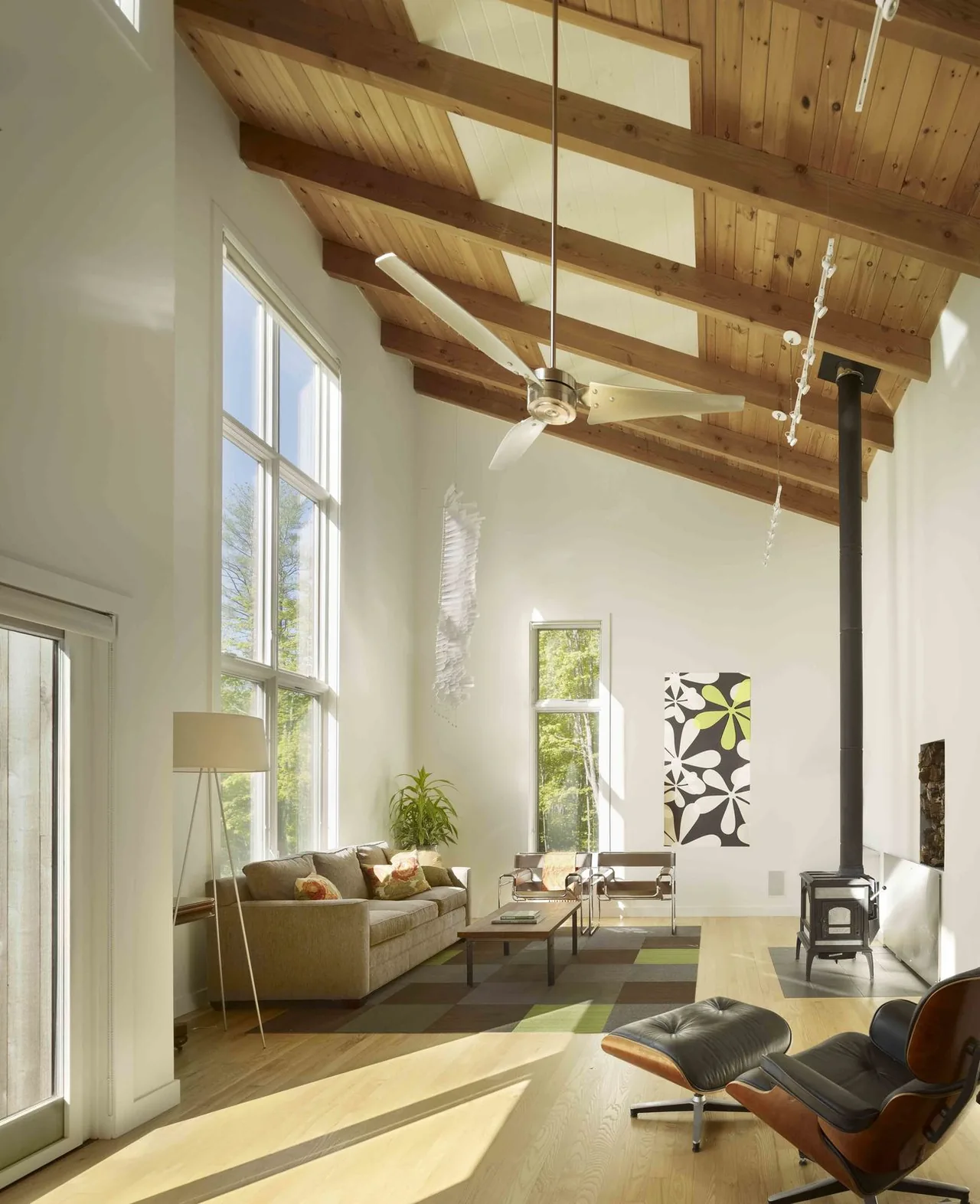 Living room interior with a vaulted ceiling with wood beams and skylights, large vertical windows, wood stove, and modern furniture.