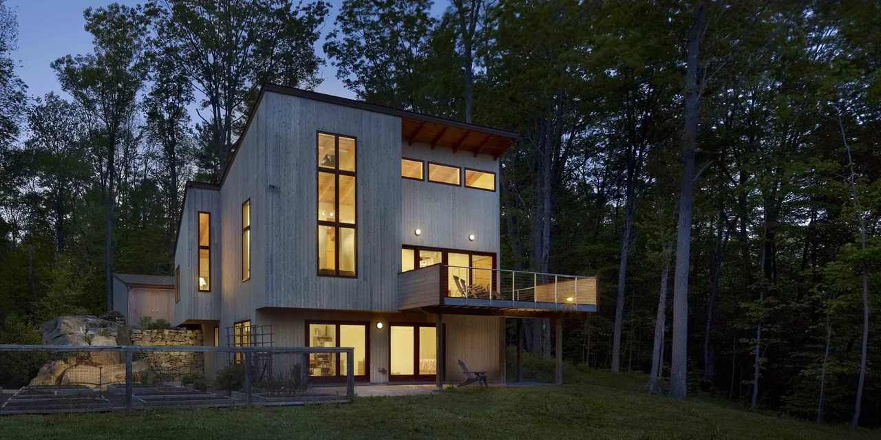 Night view of a modern flat-roofed house with wood siding, large windows, clerestory windows, and a cantilevered balcony with metal railing.