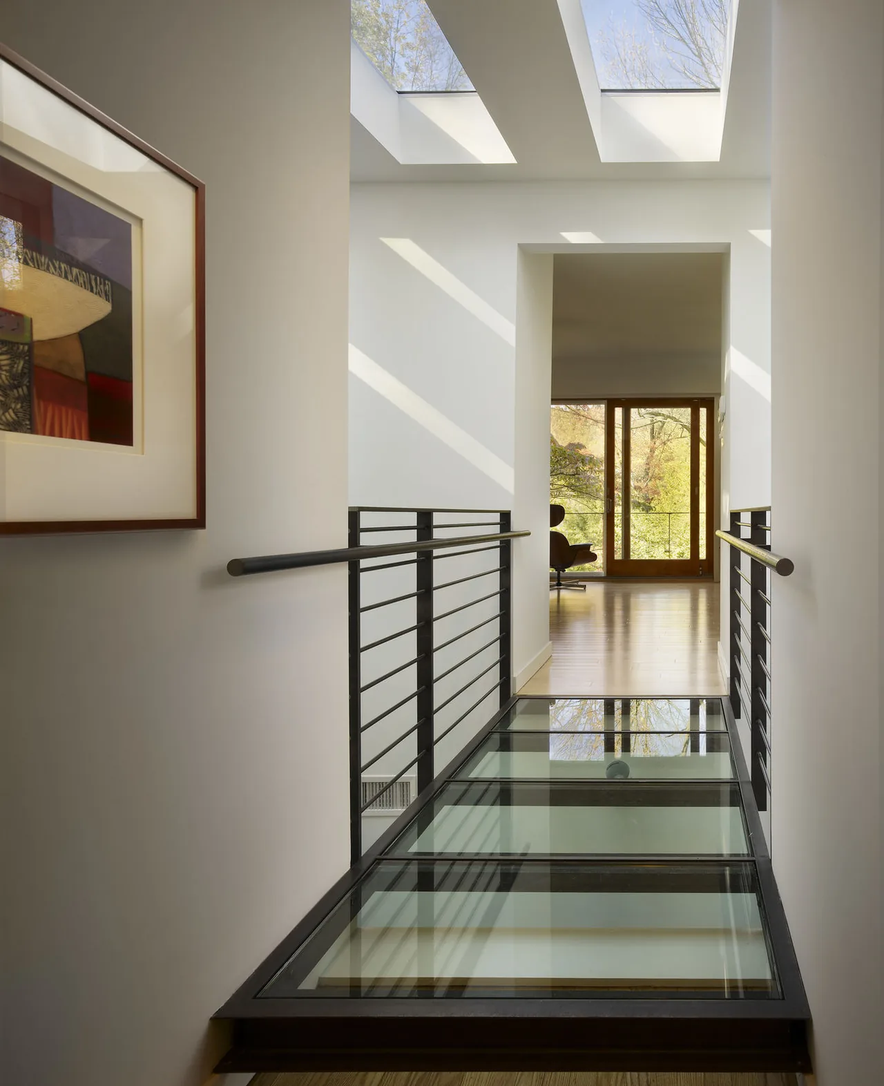 Hallway featuring glass floor panels and skylights, with metal railing and an open doorway leading to a room with wooden flooring and large windows.