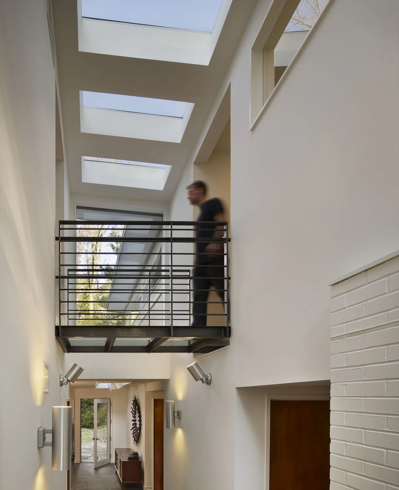 Narrow hallway illuminated by skylights and contemporary wall sconces with a metal balcony above.