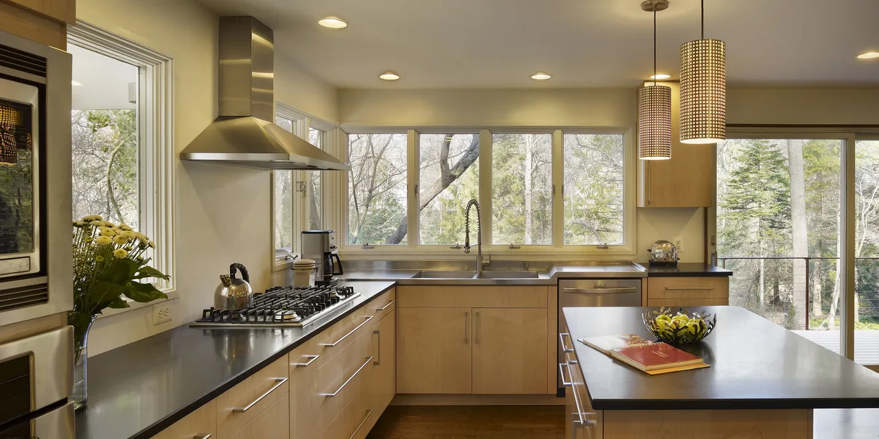 Kitchen featuring light wood cabinetry, black countertops, gas cooktop, and wide windows overlooking a wooded exterior.