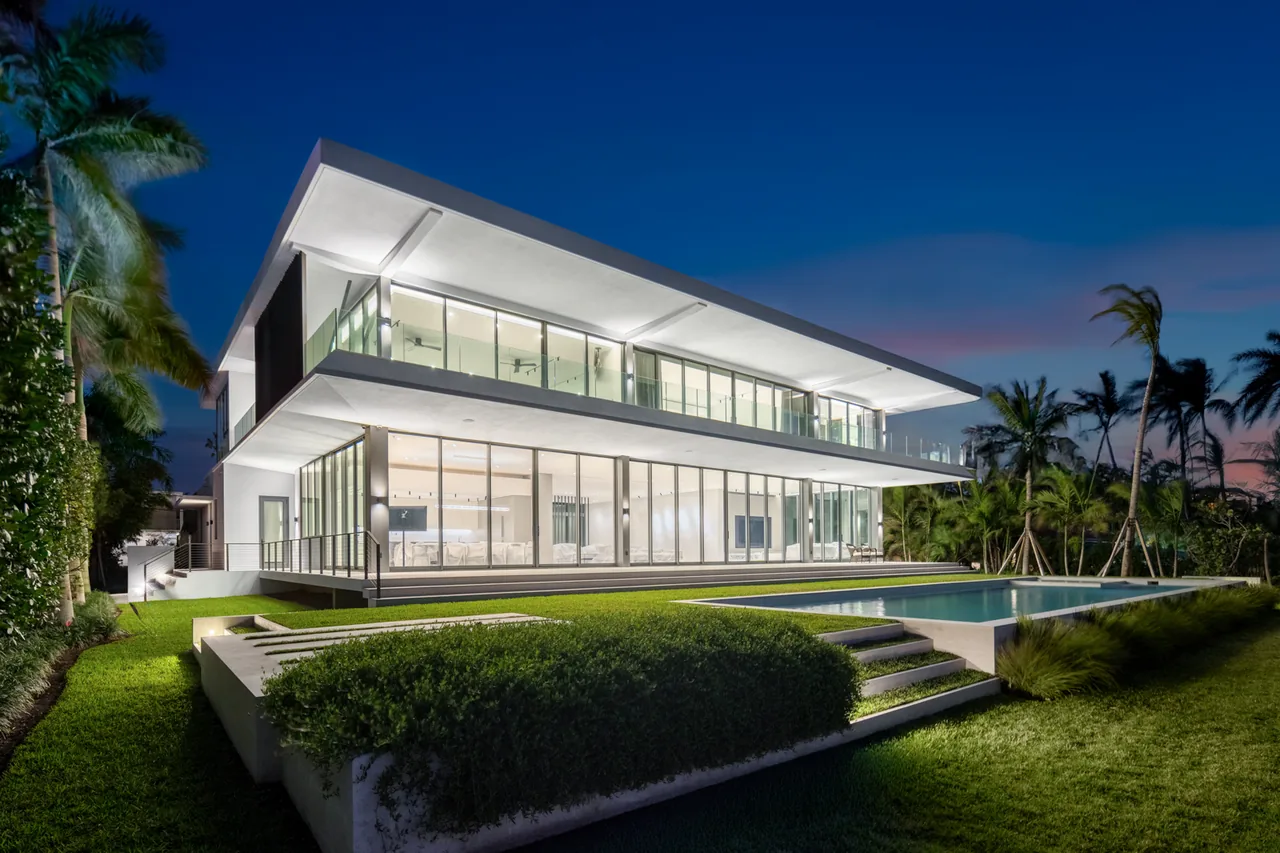 Two-story modern house at dusk with floor-to-ceiling windows, flat roof, and a pool surrounded by tropical landscaping.