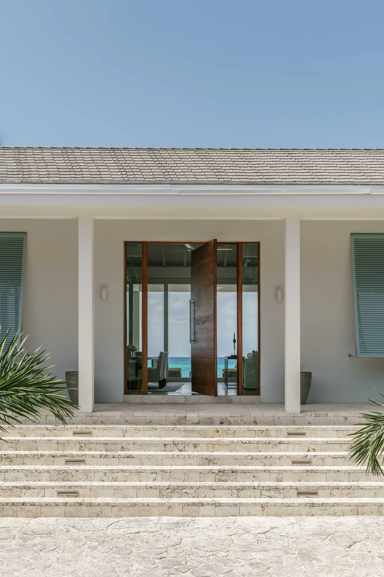 Entrance of Cat Cay Residence with stone steps, a pivot wooden door, and coastal view through glass sidelights