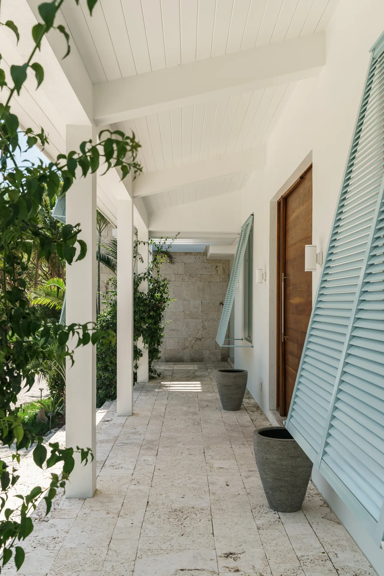 Porch at Cat Cay Residence features a vaulted white ceiling, stone tile floor, and blue louvered shutters beside a wooden door.