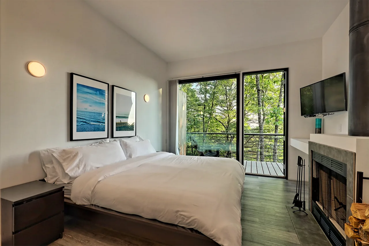 Bedroom features white bedding, large sliding glass doors to a balcony with tree views, a fireplace, and a mounted TV.