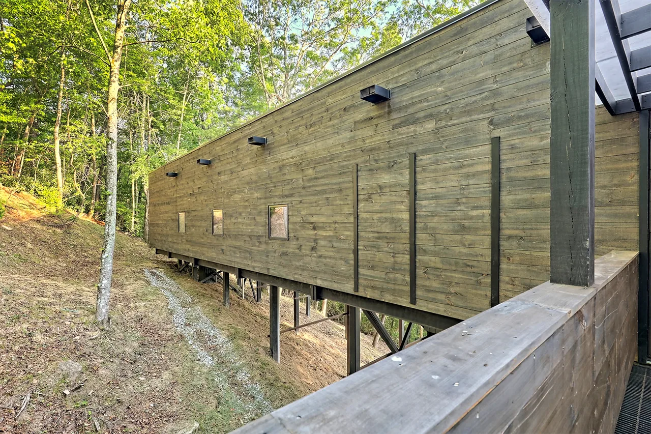 Elevated wood-sided house exterior shows structural metal supports and small windows along the length of the building.