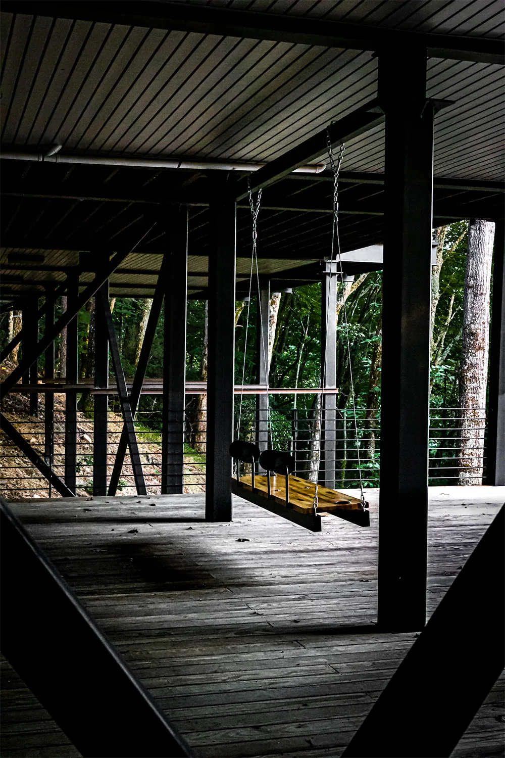 A wooden swing is suspended by chains from the ceiling on a spacious covered porch overlooking a wooded area.