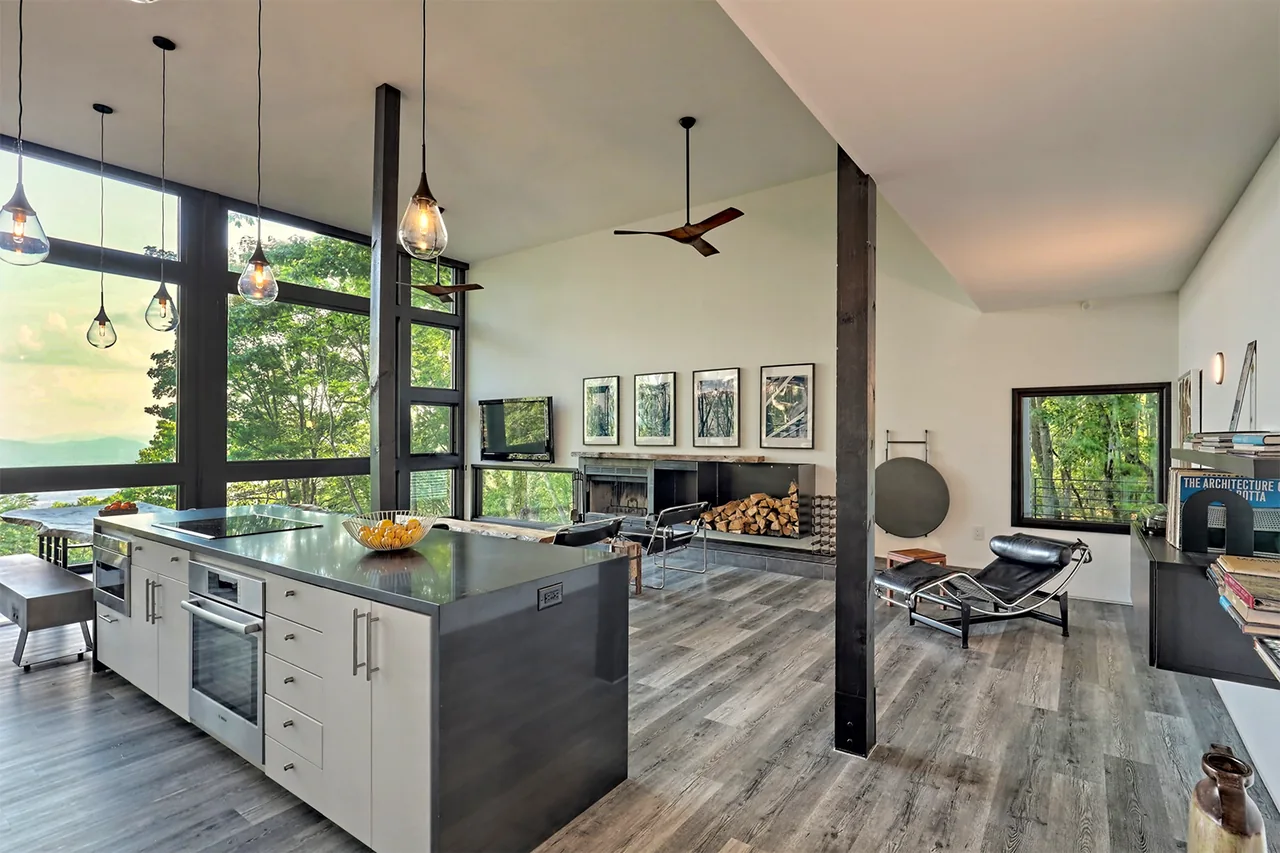 The interior includes a kitchen island, large floor-to-ceiling windows, wood flooring, and a fireplace with wood storage in the living area.