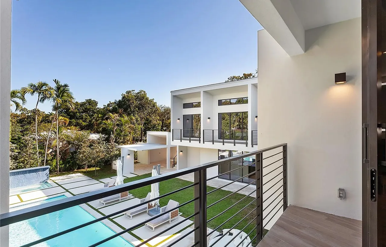 Modern house with balcony, metal railing, pool, lawn, and large windows under a flat roof.