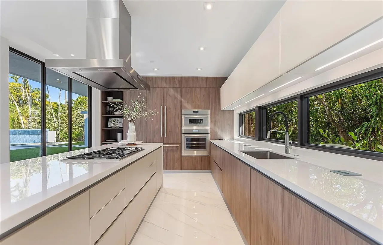 Modern kitchen features dual long counters with white countertops, wood cabinetry, and large black-framed windows providing outdoor views.