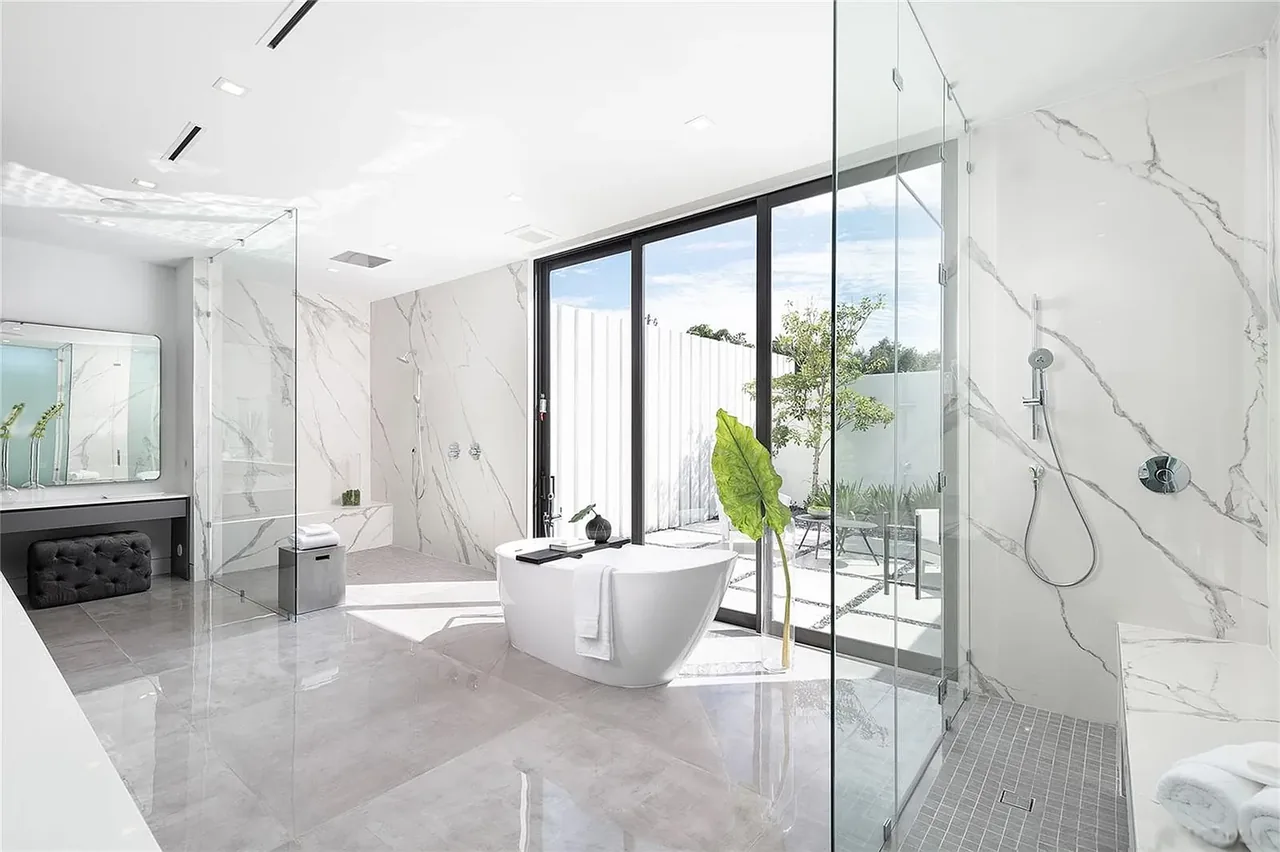The bathroom features a white freestanding tub, glass shower with marble walls, and polished floor tiles with access to an outdoor space.