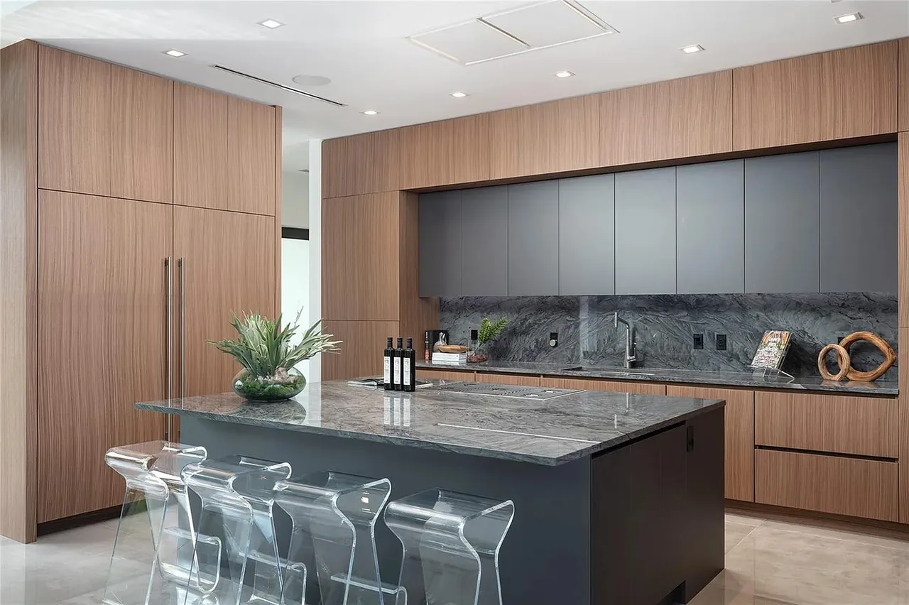 CASA BARSKY kitchen features wood-paneled lower cabinetry, smooth gray upper cabinets, and a gray stone island with transparent bar stools.