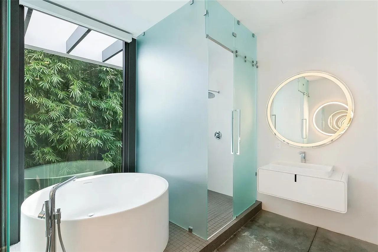 Bathroom features a white freestanding tub, frosted glass shower door, and illuminated round mirror with wall-mounted sink.