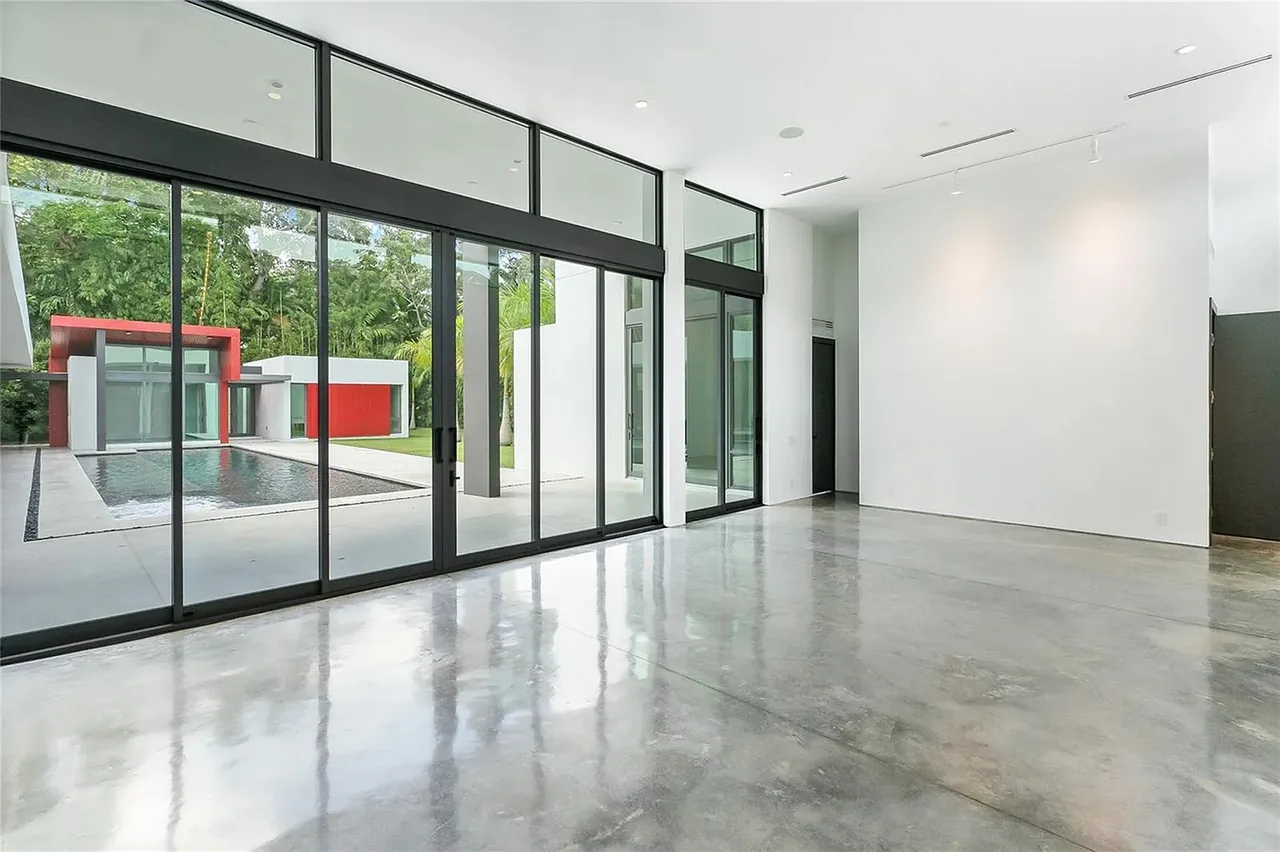 Modern living area with polished concrete flooring and floor-to-ceiling glazing overlooking pool and red-accented outdoor buildings.