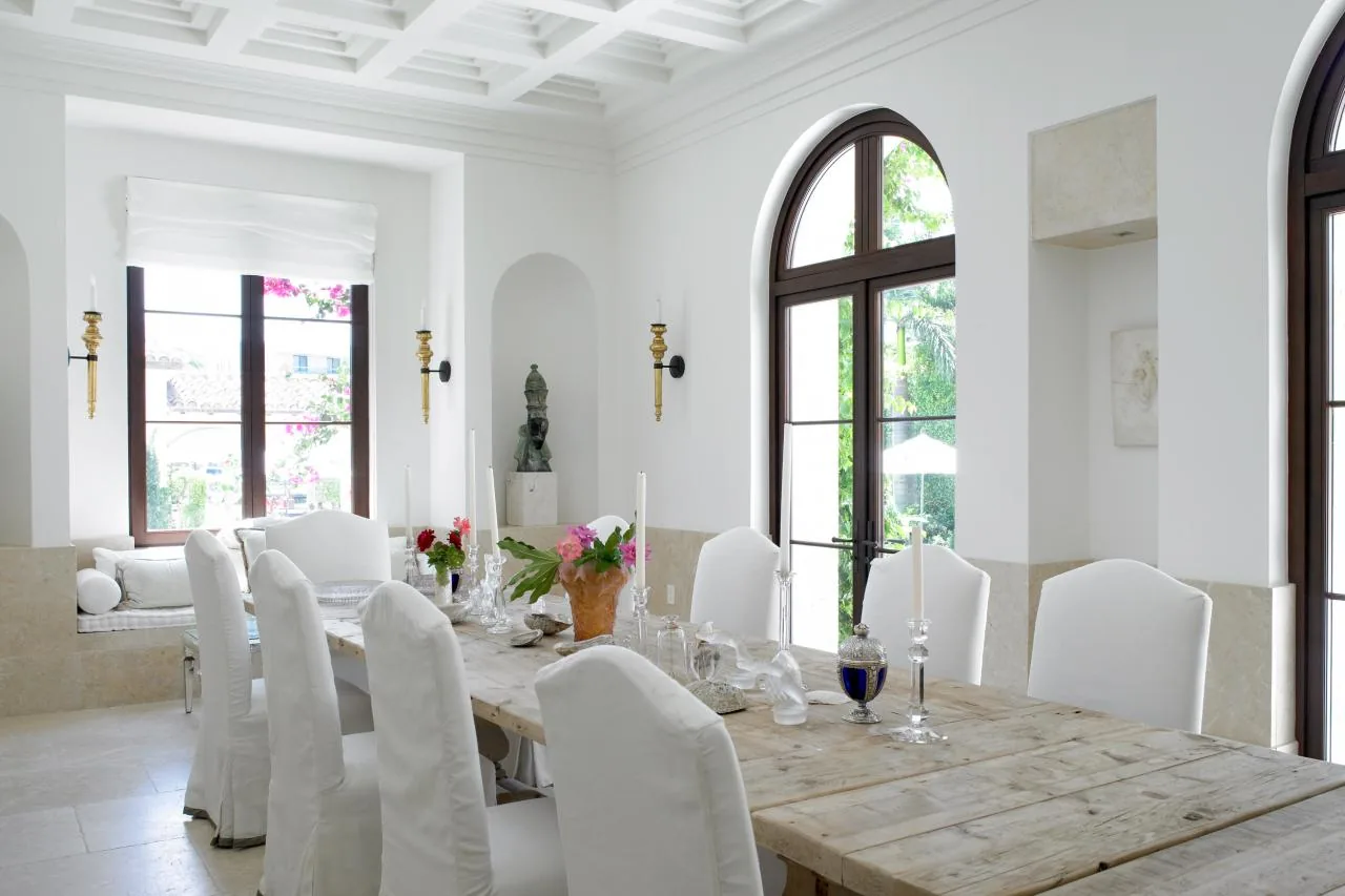 Dining room features a large natural wood table, white slipcovered chairs, arched windows with dark frames, and a coffered ceiling.