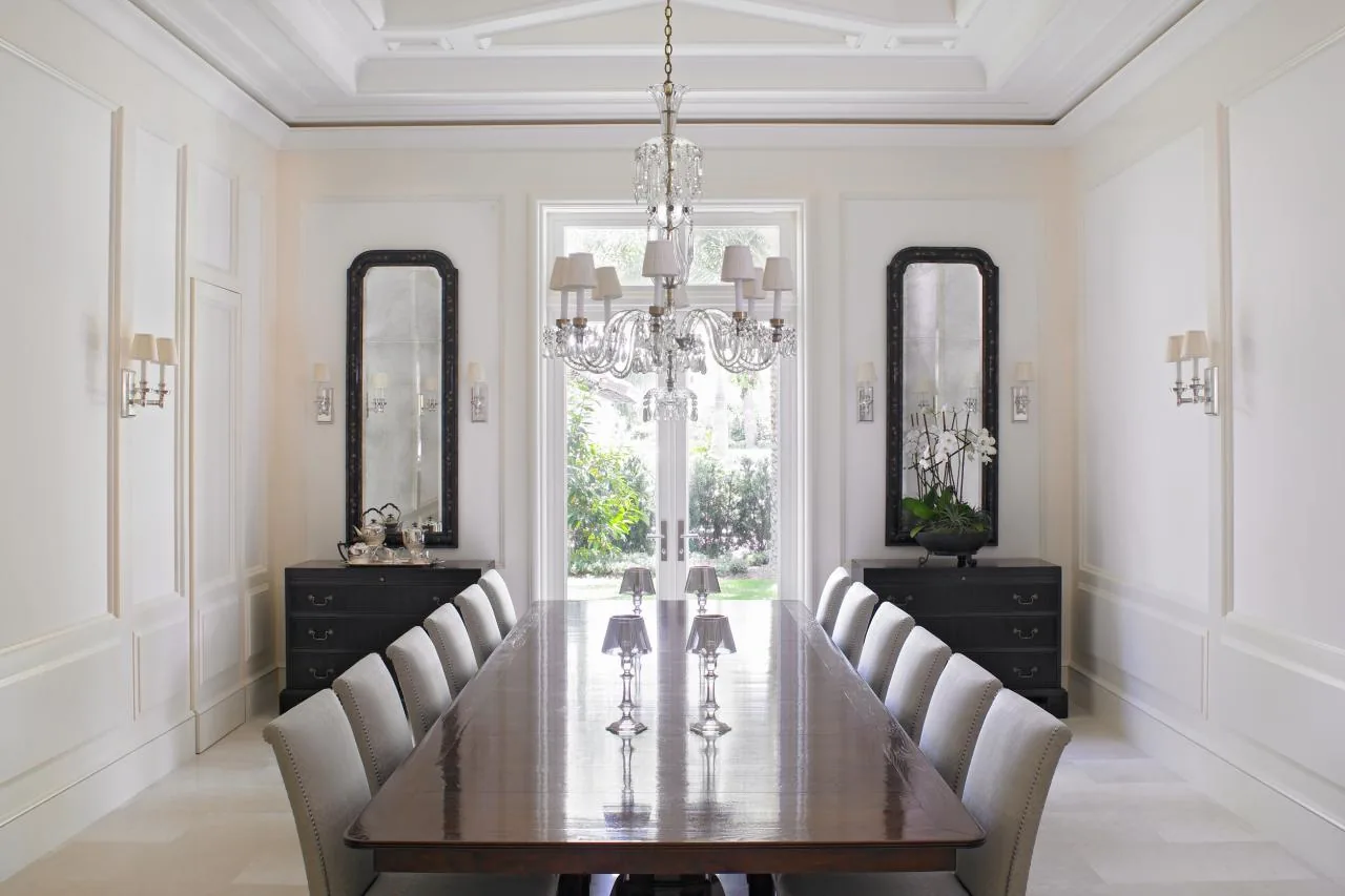 The formal dining room features a long wooden table, upholstered seating, and a crystal chandelier with natural light from glass doors.