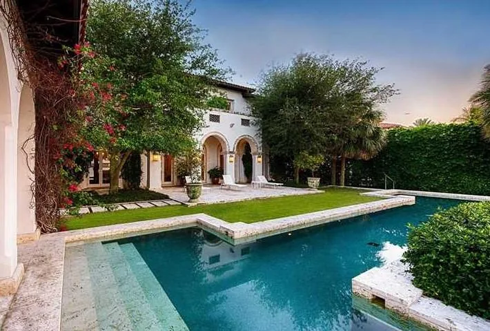 The pool area features stone coping and an arched hallway at the house facade, complemented by mature trees and hedges.