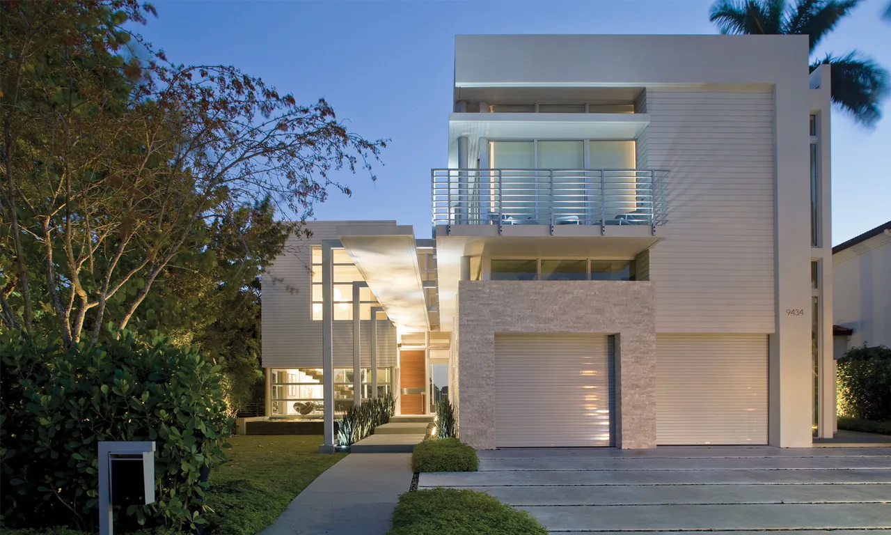 Dusk view of a modern house exterior featuring flat roofs, stone and siding walls, and metal railings.