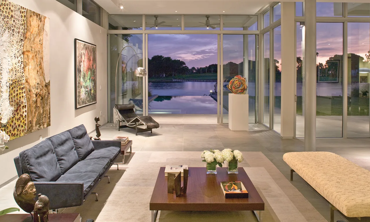 Living room with floor-to-ceiling glazing and tile flooring overlooking an outdoor pool and lake at dusk.