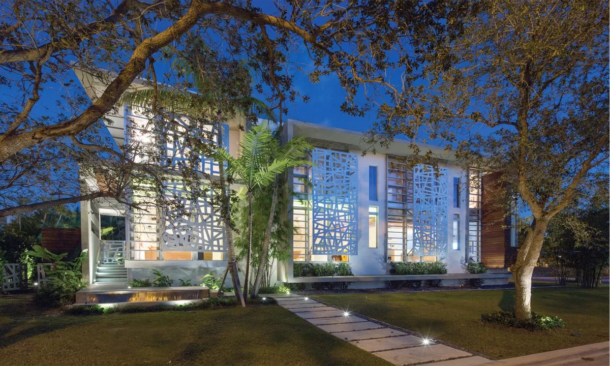 Exterior front of WOODCREST home features white decorative window screens and illuminated landscaping at twilight.