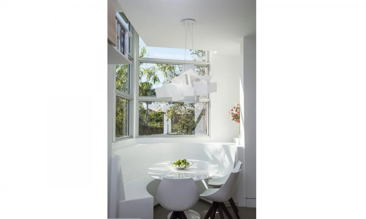 Dining nook with white built-in curved bench and round table illuminated by a geometric pendant light by large corner windows.