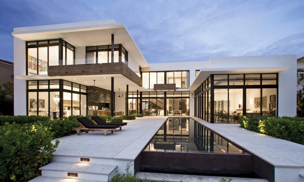 Exterior front view of a modern house featuring floor-to-ceiling glazing, flat roof, concrete and stucco materials, with a reflecting pool.