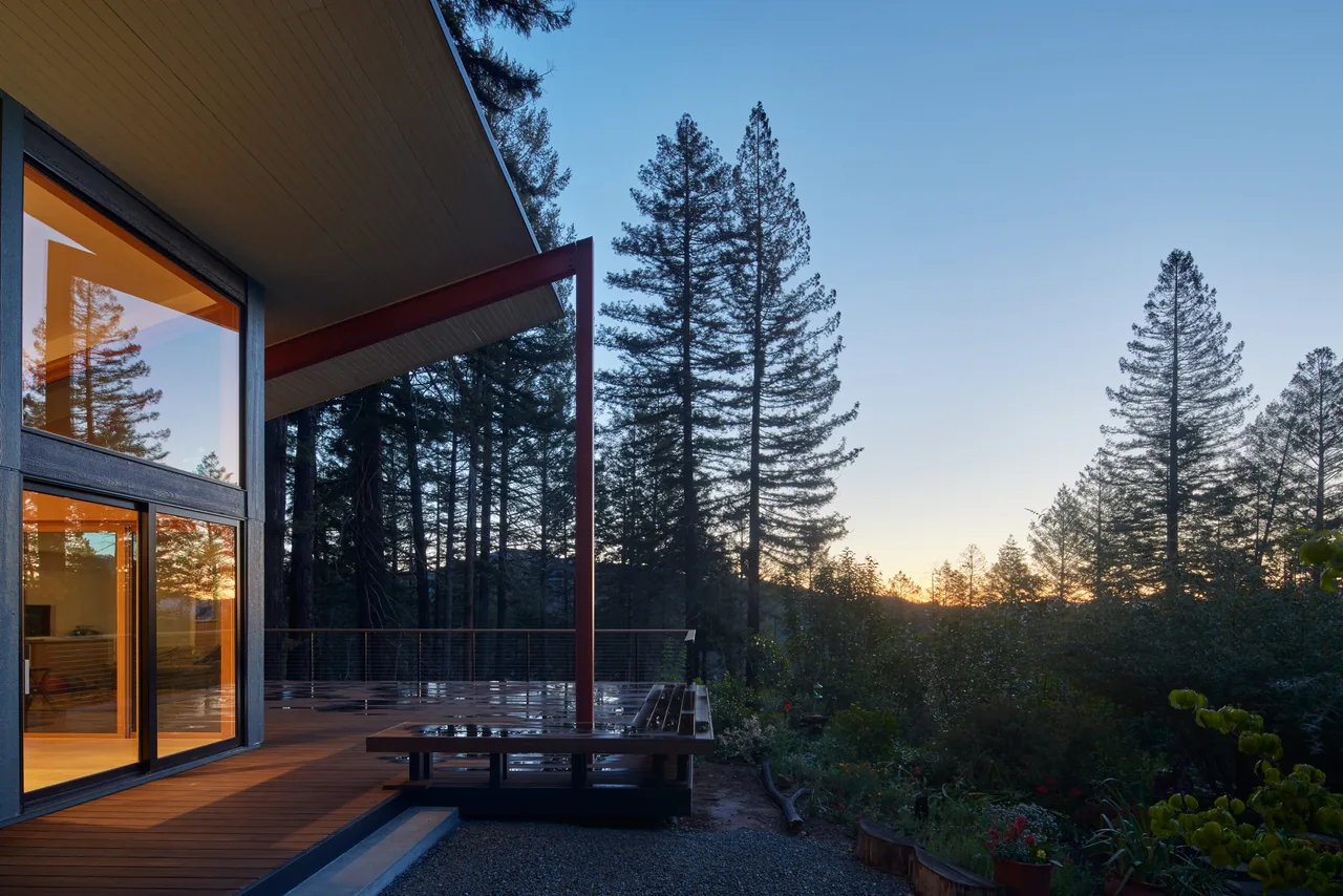 Modern house with butterfly roof and wooden deck adjacent to a forest, shown at dusk.