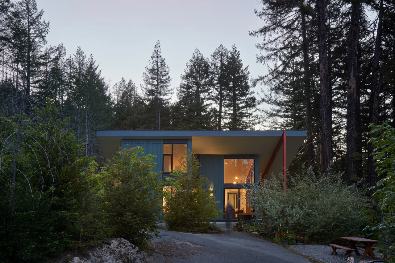 Exterior front view of a modern house with floor-to-ceiling windows and a flat roof in a wooded setting at dusk.