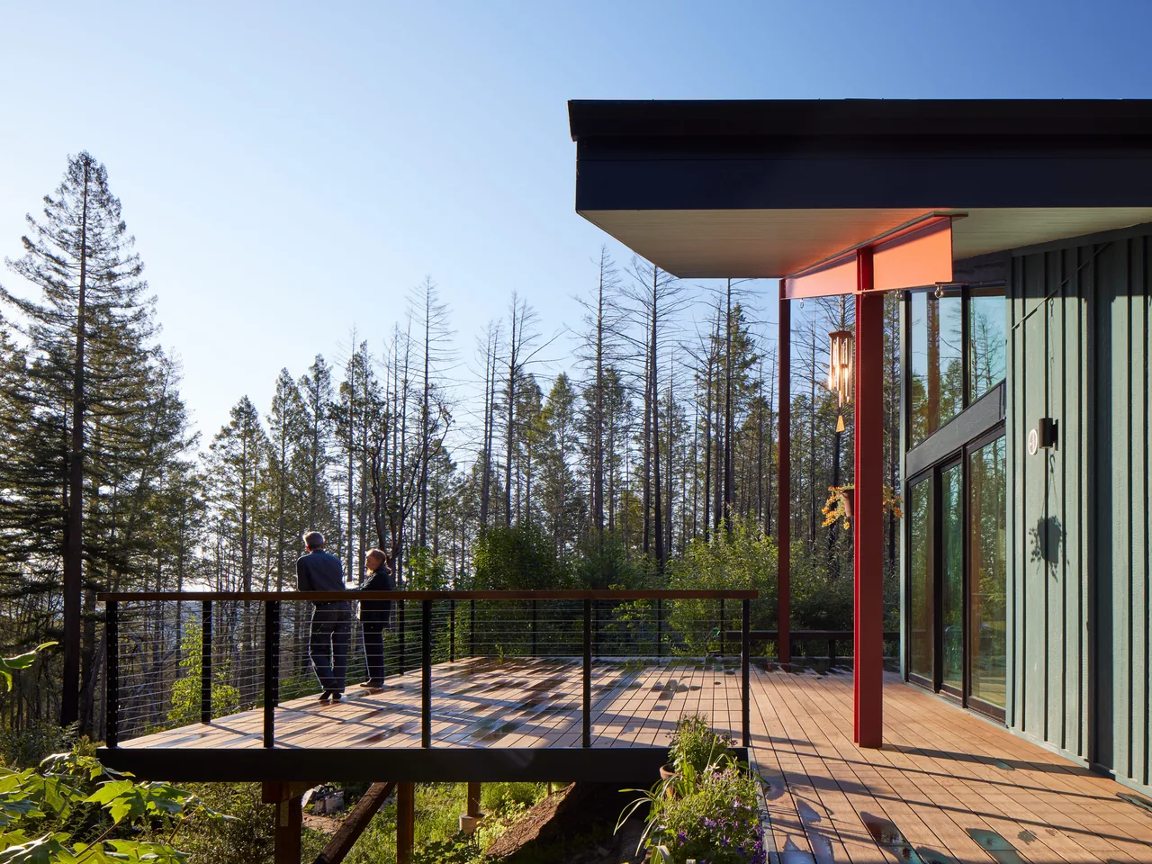 Wooden deck with black metal railing and floor-to-ceiling glazing at the side of a modern house with vertical siding.