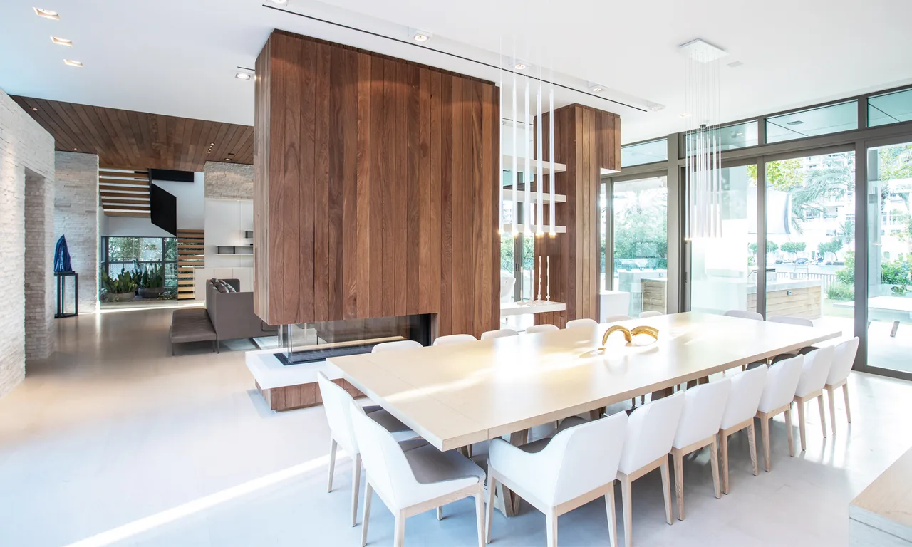 Dining room features a long table with white chairs near floor-to-ceiling glazing and a wooden partition wall with fireplace.