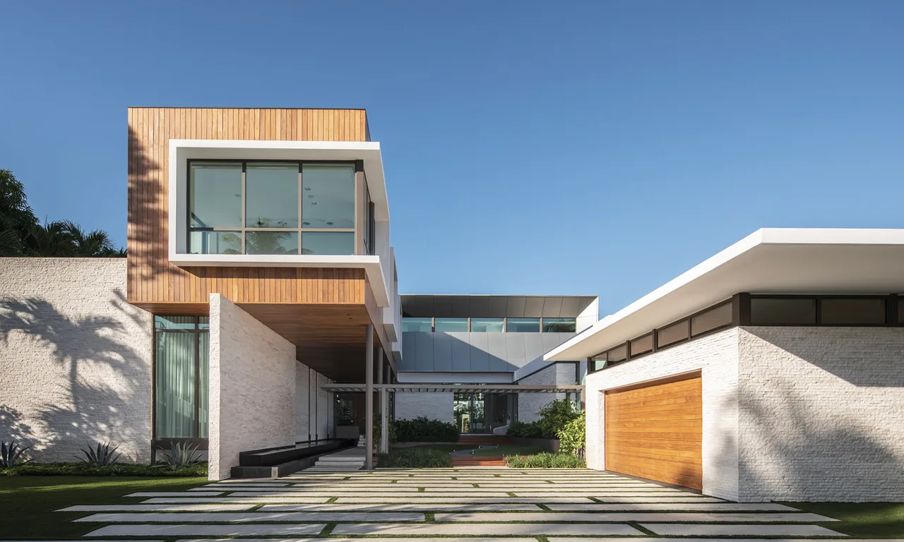 Exterior front view of a modern house featuring a wood-clad cantilevered volume, white stone walls, and a wooden garage door.