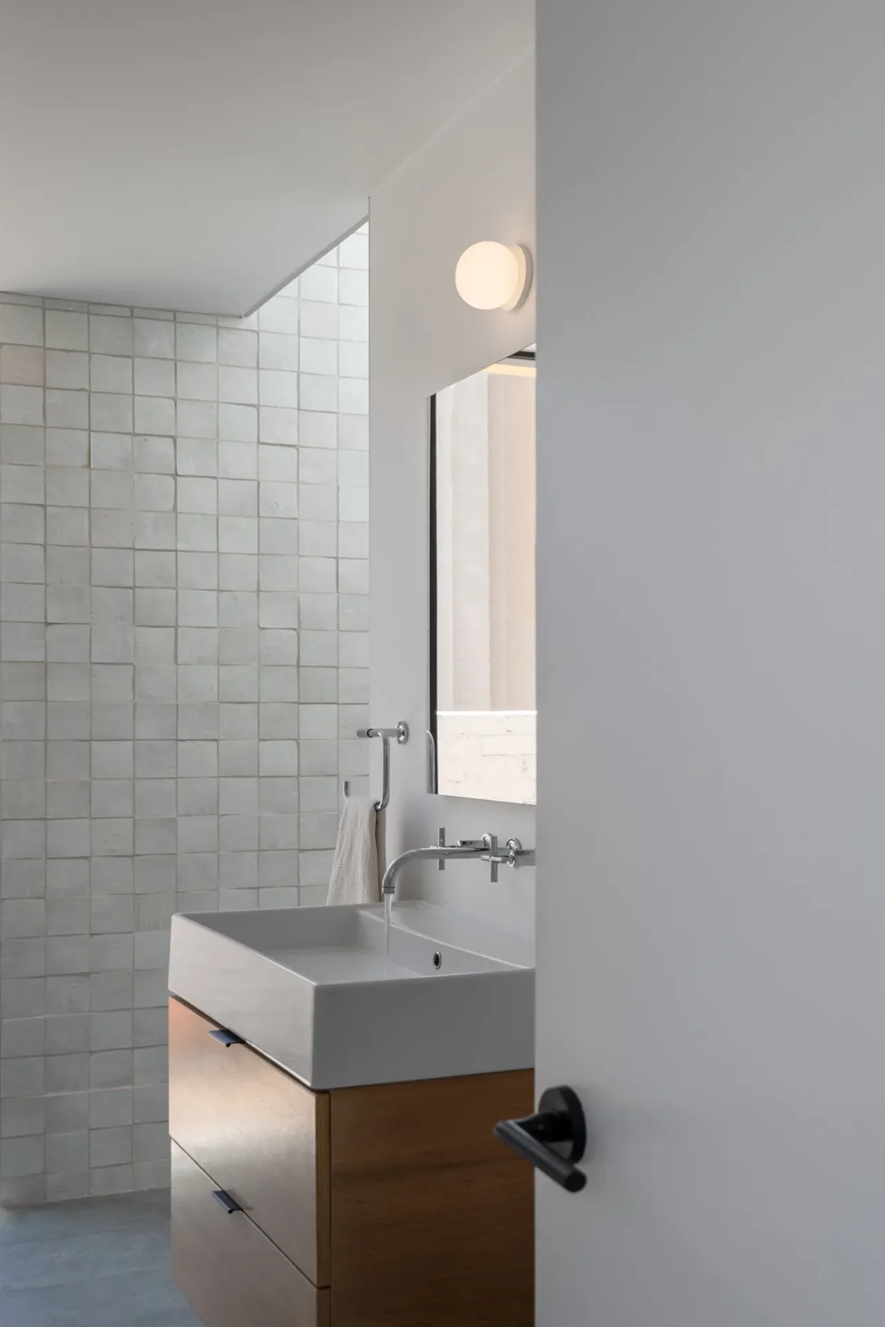 Modern bathroom interior featuring a wall-mounted faucet, square sink, and wooden vanity against a textured tiled wall.