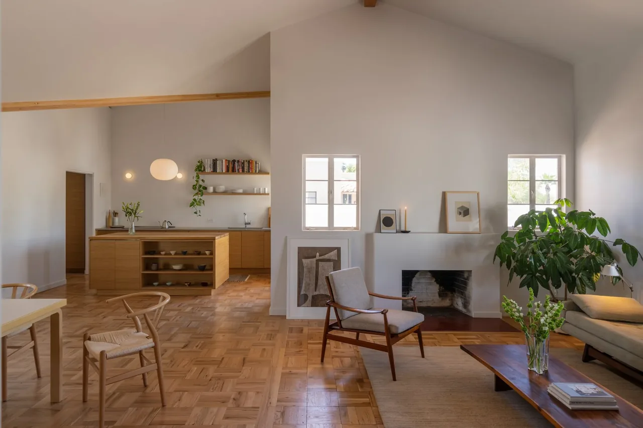 Living room and kitchen area with wood cabinetry, parquet flooring, vaulted ceiling with exposed beam, and a fireplace between two windows.