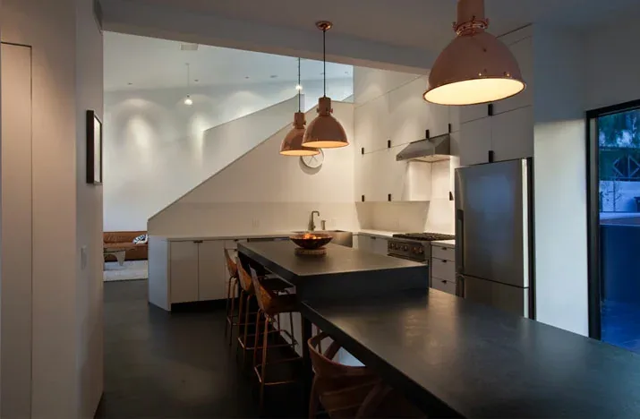 Modern kitchen interior featuring a black island with pendant lighting and a stair in the background with recessed downlights.