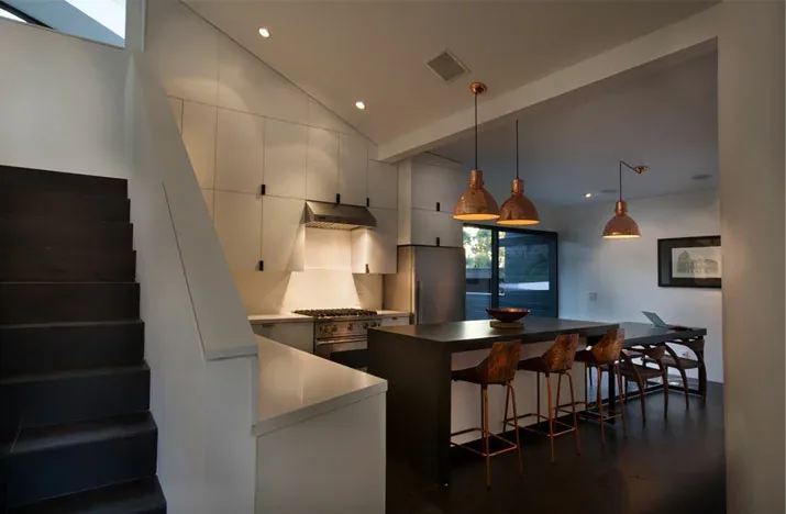 Modern kitchen and dining area with black stairs, white cabinetry, and copper pendant lighting.