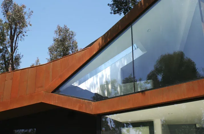 Exterior detail of a modern angular house featuring rust-colored metal cladding and large triangular windows with visible interior skylight.