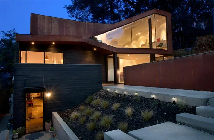 Angular modern house with rusted steel exterior cladding and extensive glass facades illuminated at dusk.