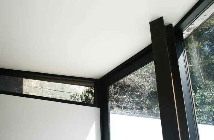 Floor-to-ceiling corner windows with black frames in a white-walled room provide natural views.