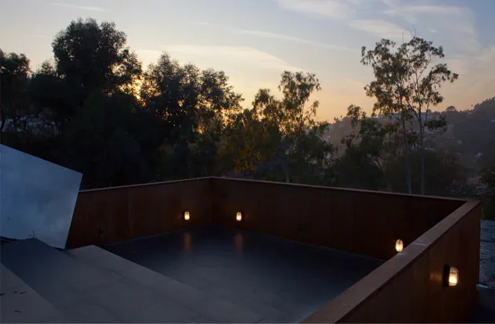 Sunken outdoor terrace with rust-colored walls and wall-mounted lights at dusk overlooking trees.