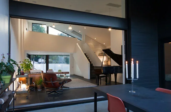 Interior view showing the living room with staircase, corner window, and floor-to-ceiling glazing opening to the patio with a dining table.