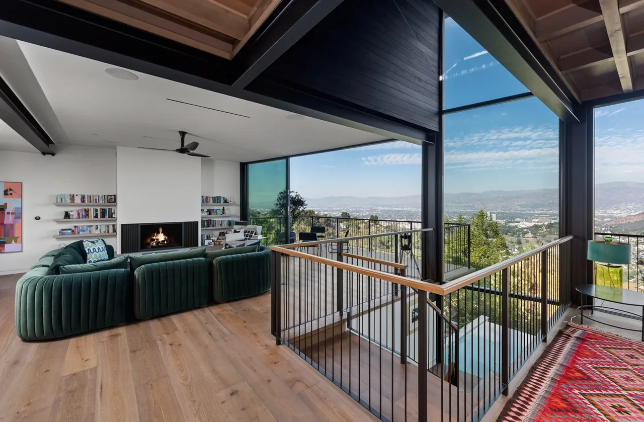 Living room with floor-to-ceiling glazing, fireplace, exposed wood ceiling beams, and metal railing next to a stairwell.