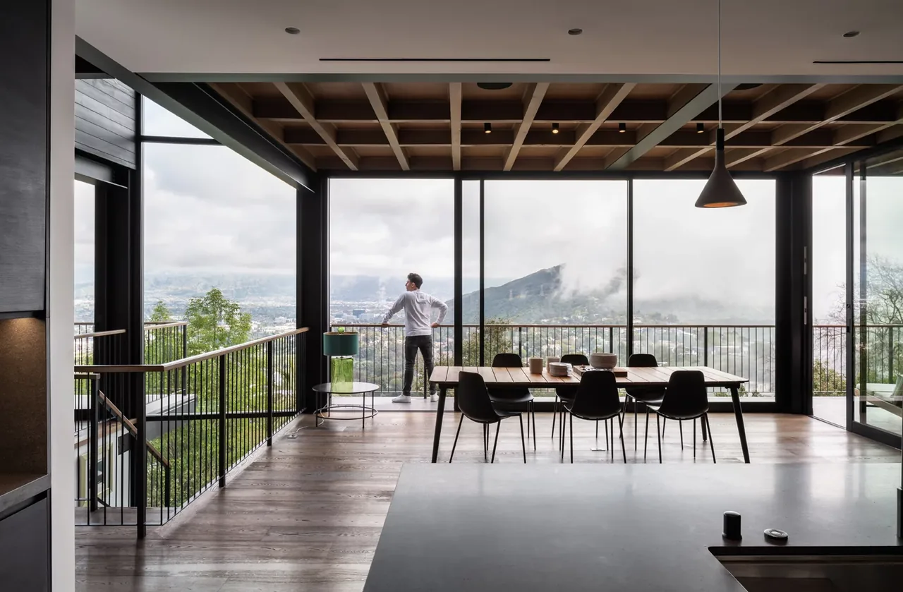 Dining area with wood ceiling beams, metal railing, and floor-to-ceiling windows overlooking a misty landscape.