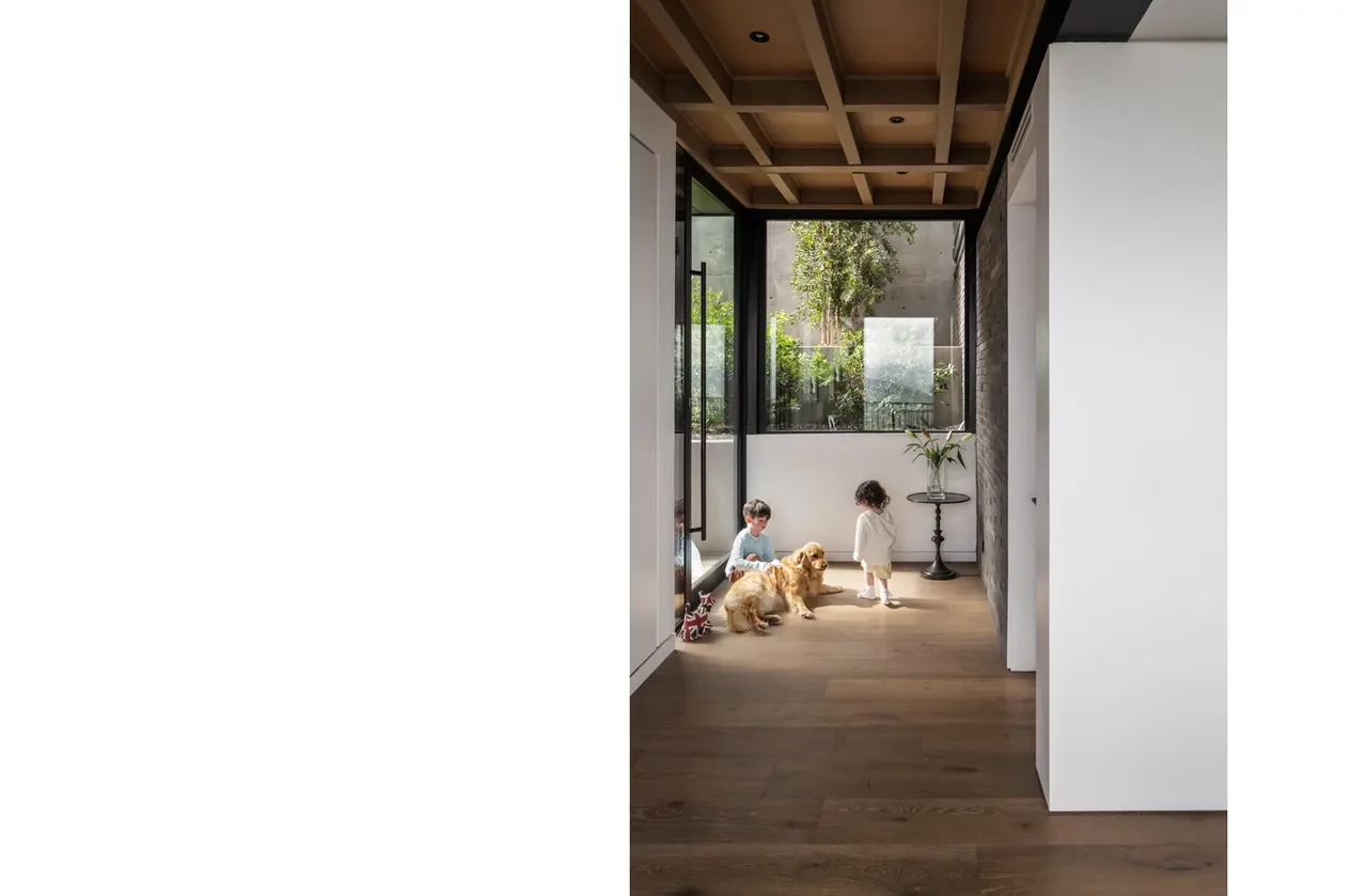 Interior corridor with wood flooring and coffered ceiling shows children and a dog near large windows.