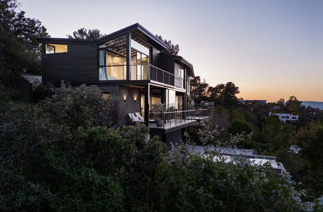 Two-story modern hillside house featuring large corner windows, balconies with metal railings, and wood siding at dusk.